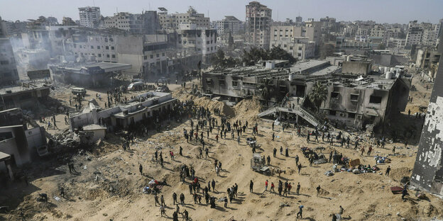 People taken from the air, standing on sandy ground, with debris from high-rise buildings in the background