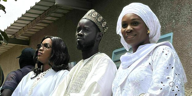 The new Senegalese president, Bassirou Diomaye Faye, stands between his two wives, all three dressed in white.