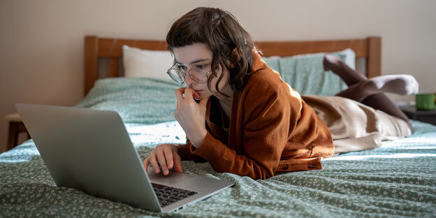 A teenager lies on a bed and looks with interest at a laptop.