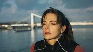 "Bauchgefühl - Lena": Lena (Laura Berlin) steht hoch auf einer Brücke. Im Hintergrund ist eine weitere Brücke zu sehen, unter ihr fährt ein Frachtschiff auf dem breiten Fluss entlang.