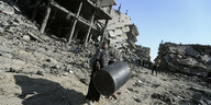 Eine Frau trägt eine Tonne durch ein zerstörtes Gebiet mit kaputten Häusern