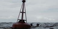 Eine Szene auf der Ostsee: Zwei Menschen in Tauscheranzügen und mit Taucherbrillen schwimmen auf eine rote Metallboje zu, die auf dem Meer aufsitzt. Der Himmel und das Wasser sind grau.