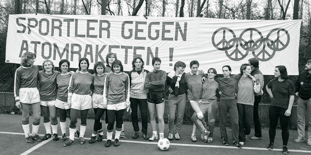 Schwarz-weiß-Fotografie. Eine Gruppe Frauen in Sportklamotten und mit einem Fußball stehen nebeneinander vor einem großen Banner, auf dem steht: "Sportler gegen Atomraketen"