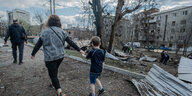 Eine Frau hält ein Kind an ihrer Hand, beide laufen an zerstörten Gebäuden vorbei