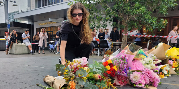 A young woman with a tearful face and sunglasses places flowers on the street