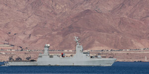 Ein graues Kriegsschiff fährt auf Wasser, im Hintergrund eine sandige Landschaft