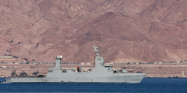 A gray warship sails on the water, with a sandy landscape in the background