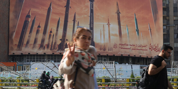 A woman stands in front of a poster with rockets and makes a peace sign with her hand.