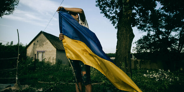 A young man stands in the front yard of a small village in the Kharkiv region and raises a Ukrainian flag.