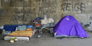 Ein Matratzenlager, Einkaufswagen gefüllt mit Sachen, ein Zelt vor einer Mauer