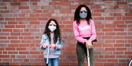 Zwei Mädchen stehen mit Stoffmasken vor einer Backsteinwand - sie sind mit Rollern unterwegs