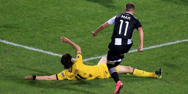 Ein Fußballspieler liegt quer auf dem Rasen, hinter ihm ein anderer Spieler, der gerade den Ball gespielt hat Ein Fußballspieler liegt quer auf dem Rasen, hinter ihm ein anderer Spieler, der gerade den Ball gespielt hat