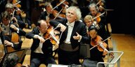 Blick auf ein Orchester. Der Dirigent Simon Rattle steht am Dirigentenpult und bewegt den Taktstock und seine Hand zur Musik. Er lächelt.