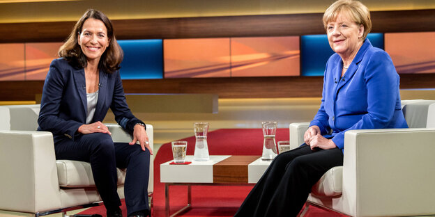 Eine dunkelhaarige Frau sitzt in einem Sessel, neben ihr auf einem Sessel Angela Merkel Eine dunkelhaarige Frau sitzt in einem Sessel, neben ihr auf einem Sessel Angela Merkel