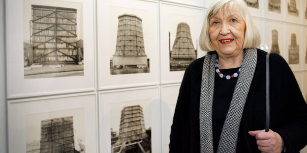 Eine Frau mit grauen Haaren steht vor einer Wand mit Fotografien von Kühltürmen Eine Frau mit grauen Haaren steht vor einer Wand mit Fotografien von Kühltürmen