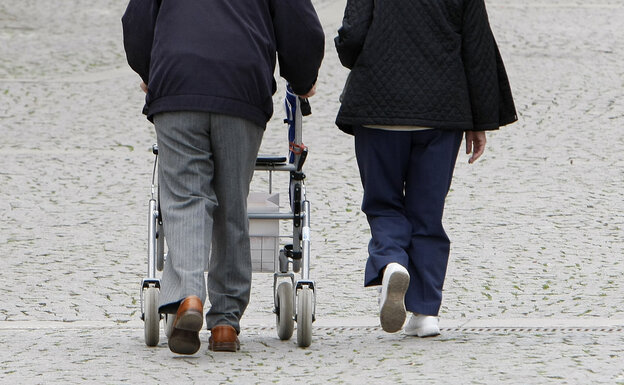 Ein alter Mensch mit einem Rollator läuft neben einem anderen Menschen auf Kopfsteinpflaster entlang.