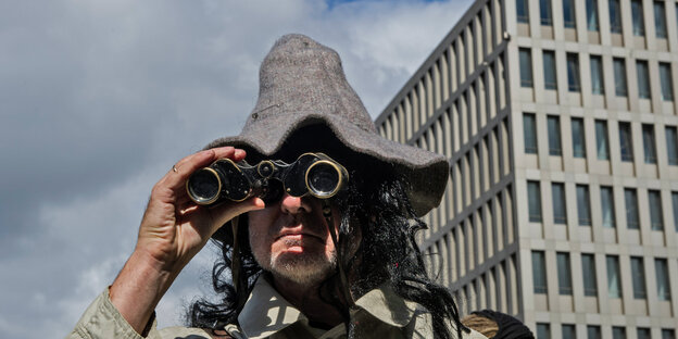 Mann mit Schlapphut und Fernglas vor modernem Bürogebäude Mann mit Schlapphut und Fernglas vor modernem Bürogebäude