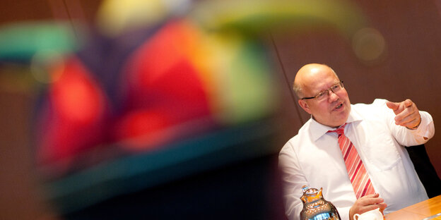 Unscharf im Vordergrund eine bunte Mütze, hinten am Tisch sitzt ein kahler Mann mit Krawatte, es ist Kanzleramtsminister Peter Altmaier (CDU), der auch für Geheimdienste zuständig ist Unscharf im Vordergrund eine bunte Mütze, hinten am Tisch sitzt ein kahler Mann mit Krawatte, es ist Kanzleramtsminister Peter Altmaier (CDU), der auch für Geheimdienste zuständig ist