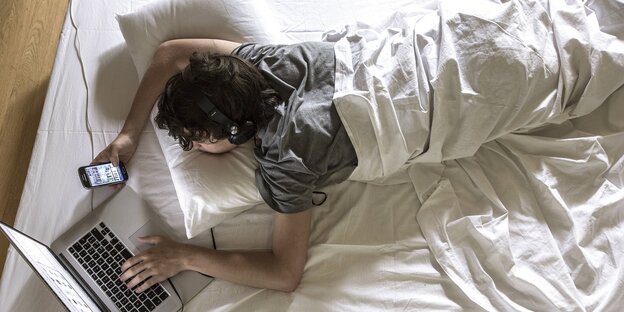 Ein junger Mann liegt mit Laptop und Smartphone im Bett Ein junger Mann liegt mit Laptop und Smartphone im Bett