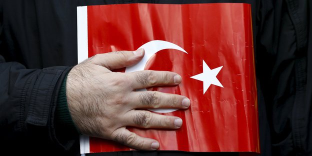 Ein türkisches Fähnchen wird von einer behaarten Hand gegen eine unbekannte Brust gedrückt.