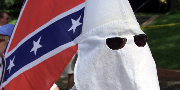 Ein Mitglied des Ku-Klux Klans mit seiner weißen Mütze und Sonnenbrille. Im Hintergrund die konföderierten Flagge Ein Mitglied des Ku-Klux Klans mit seiner weißen Mütze und Sonnenbrille. Im Hintergrund die konföderierten Flagge