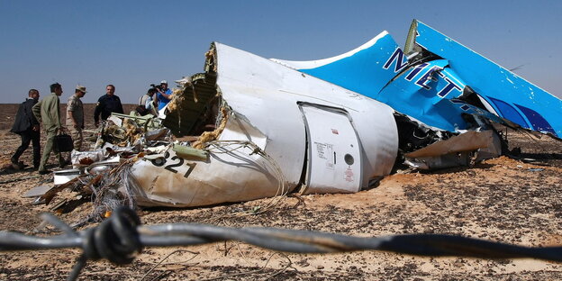 Wrackteile des in Ägypten abgeschossenen russischen Flugzeugs. Im Hintergrund sind ein paar Menschen vor blauem Himmel zu sehen, die die Absturzstelle untersuchen. Vor der Kameralinse verläuft unscharf Stacheldrahtzaun. Wrackteile des in Ägypten abgeschossenen russischen Flugzeugs. Im Hintergrund sind ein paar Menschen vor blauem Himmel zu sehen, die die Absturzstelle untersuchen. Vor der Kameralinse verläuft unscharf Stacheldrahtzaun.