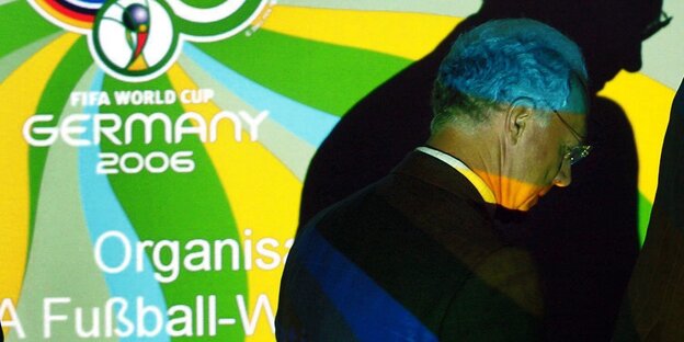 Franz Beckenbauer steht neben einem Plakat zur WM 2006. Sein Kopf wirft einen dunklen Schatten auf das Plakat. Franz Beckenbauer steht neben einem Plakat zur WM 2006. Sein Kopf wirft einen dunklen Schatten auf das Plakat.