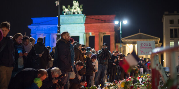 Menschen stehen an einer Streifen aus Blumen und Kerzen. Im Hintergund das Brandenburger Tor in blau, weiß, rot. Menschen stehen an einer Streifen aus Blumen und Kerzen. Im Hintergund das Brandenburger Tor in blau, weiß, rot.