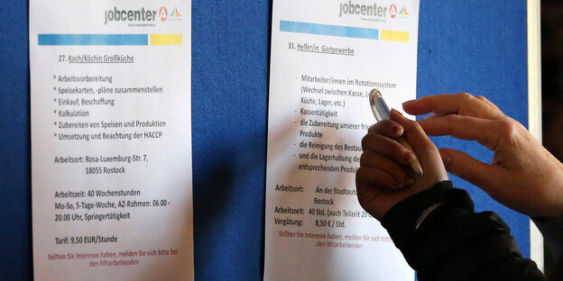 An einem Aufsteller hängen Zettel mit Jobangeboten, zwei Hände zeigen auf einen der Zettel An einem Aufsteller hängen Zettel mit Jobangeboten, zwei Hände zeigen auf einen der Zettel