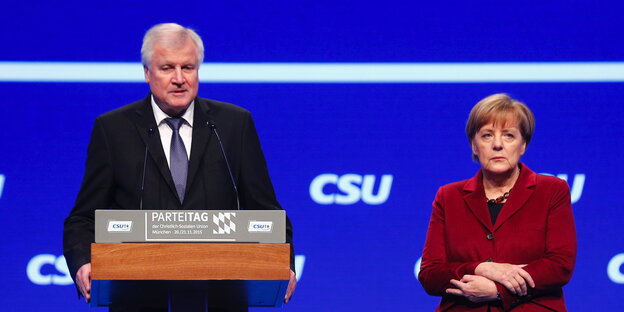 Horst Seehofer tront an seinem Rednerpult, Frau Merkel steht bedröppelt daneben Horst Seehofer tront an seinem Rednerpult, Frau Merkel steht bedröppelt daneben