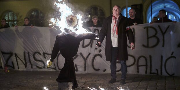 Im Vordergrund brennt eine Puppe, dahinter steht ein Mann mit einer weiß-roten Armbinde und einer Flasche Brandbeschleuniger in der Hand. Im Hintergrund stehen rechtsradikale Demonstranten hinter einem Banner mit faschistischen Parolen auf polnisch.