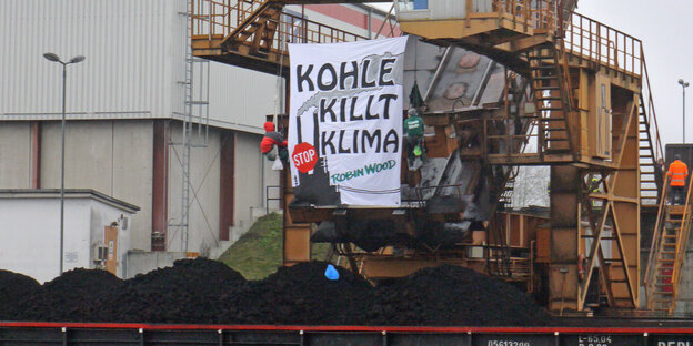 Kran mit Kohlehaufen davor, vor dem Steuerhäuschen hängt ein Plakat mit der Aufschrift "Kohle killt Klima", daneben zwei angeseilte Menschen Kran mit Kohlehaufen davor, vor dem Steuerhäuschen hängt ein Plakat mit der Aufschrift "Kohle killt Klima", daneben zwei angeseilte Menschen