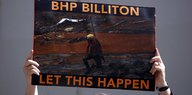 mensch hält Plakat hoch, auf dem steht (deutsche Übersetzung): "BHP Billiton hat das zugelassen", auf dem Plakat ein roter Fluss zu sehen, alle Vegetation drumrum tot