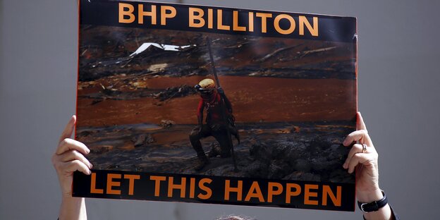 mensch hält Plakat hoch, auf dem steht (deutsche Übersetzung): "BHP Billiton hat das zugelassen", auf dem Plakat ein roter Fluss zu sehen, alle Vegetation drumrum tot mensch hält Plakat hoch, auf dem steht (deutsche Übersetzung): "BHP Billiton hat das zugelassen", auf dem Plakat ein roter Fluss zu sehen, alle Vegetation drumrum tot