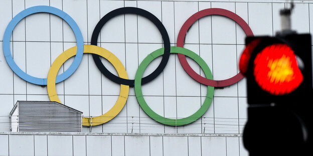 Die fünf Olympischen Ringe an einer Hauswand hinter einen roten Ampel Die fünf Olympischen Ringe an einer Hauswand hinter einen roten Ampel