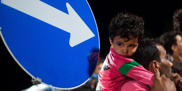 Schild mit Pfeil, der auf ein Flüchtlingskind weist, dass auf der Schulter seines Vaters sitzt. Schild mit Pfeil, der auf ein Flüchtlingskind weist, dass auf der Schulter seines Vaters sitzt.