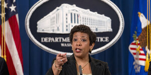 Frau vor einem ovalen Enblem, auf dem "Department of Justice" steht, links und rechts eine US-Fahne