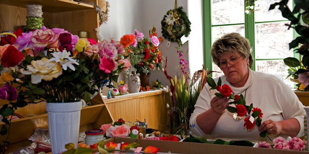 Eine Frau fertigt Kunstblumen, neben sich ein großer, bunter Straus in einem Eimer Eine Frau fertigt Kunstblumen, neben sich ein großer, bunter Straus in einem Eimer