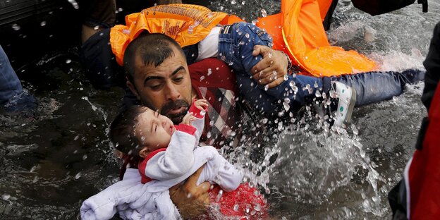 Ein Mann mit Schwimmweste steht im Wasser und hält ein Baby und ein Kleinkind im Arm Ein Mann mit Schwimmweste steht im Wasser und hält ein Baby und ein Kleinkind im Arm