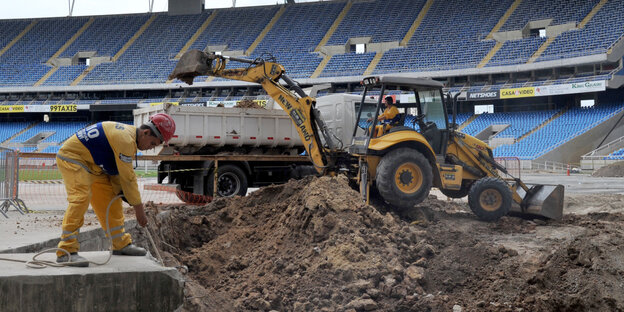 Ein Bagger und ein Bauarbeiter stehen mitten in einem unfertigen Stadion Ein Bagger und ein Bauarbeiter stehen mitten in einem unfertigen Stadion