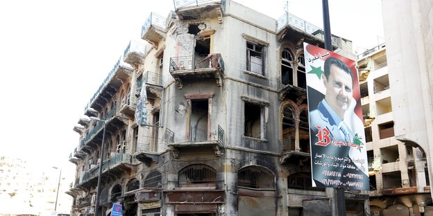 Baschar al-Assad blickt von einem Plakat herab, das an einer Straßenlaterne angebracht ist - im Hintergrund ein zerstörtes Gebäude. Baschar al-Assad blickt von einem Plakat herab, das an einer Straßenlaterne angebracht ist - im Hintergrund ein zerstörtes Gebäude.