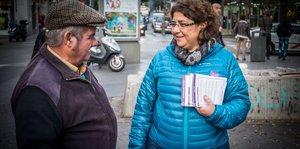 Eine Frau hält Infomaterial in der Hand und spricht auf der Straße mit einem Mann.