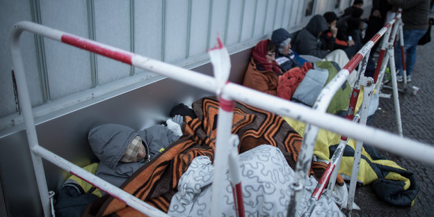 Flüchtlinge sitzen auf einem Bürgersteig und sind in Decken eingehüllt.