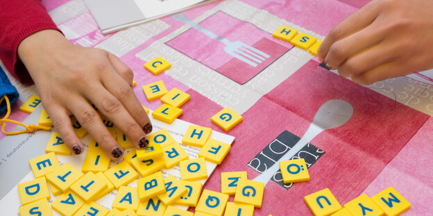 Hände schieben gelbe Plättchen mit Buchstaben über eine rot-weiße Tischdecke Hände schieben gelbe Plättchen mit Buchstaben über eine rot-weiße Tischdecke