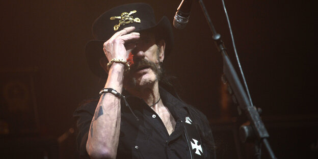 Lemmy Kilmister steht hinter einem Mikro und hält die Hand an seinen Hut Lemmy Kilmister steht hinter einem Mikro und hält die Hand an seinen Hut