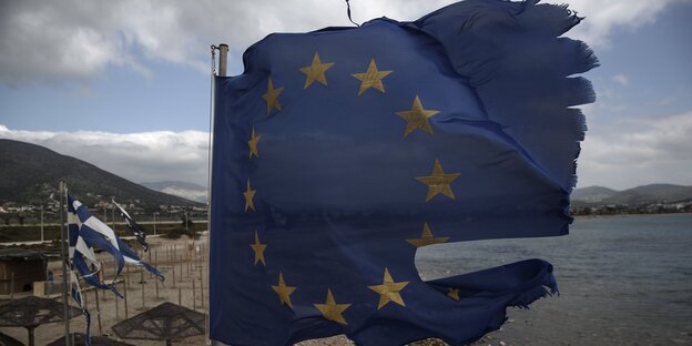Ruinierte EU und griechische Flaggen fliegen in Fetzen von einem Fahnenmast auf einem Strand. Ruinierte EU und griechische Flaggen fliegen in Fetzen von einem Fahnenmast auf einem Strand.