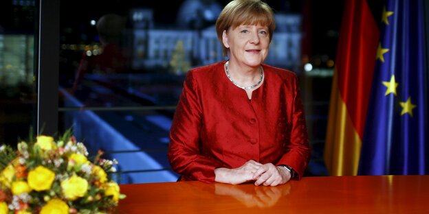 Angela Merkel steht in einem roten Blazer an einem Tisch, auf ihm stehen gelbe Blumen Angela Merkel steht in einem roten Blazer an einem Tisch, auf ihm stehen gelbe Blumen