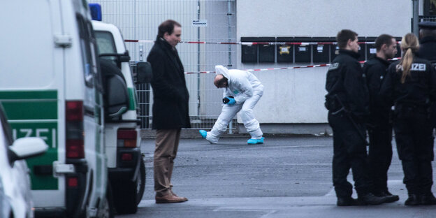 Polizisten stehen auf einem Parkplatz herum, im Hintergrund ein Mann im weißen Overall bei der Sicherung von Spuren. Polizisten stehen auf einem Parkplatz herum, im Hintergrund ein Mann im weißen Overall bei der Sicherung von Spuren.