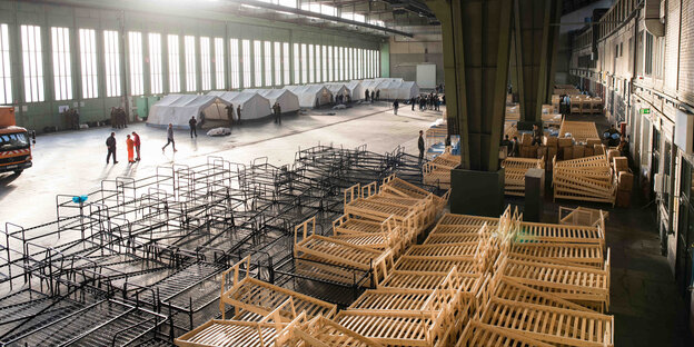 Dutzende Betten stehen teils gestapelt in einem Hangar des ehemaligen Flughafens Tempelhof in Berlin. Dutzende Betten stehen teils gestapelt in einem Hangar des ehemaligen Flughafens Tempelhof in Berlin.