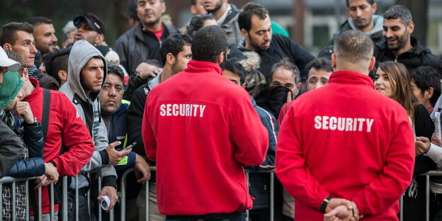 zwei Security-Mitarbeiter stehen vor einer Absperrung vor vielen Flüchtlingen zwei Security-Mitarbeiter stehen vor einer Absperrung vor vielen Flüchtlingen